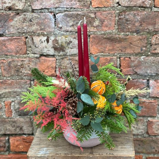 Mulled Spice Christmas table arrangement with red candles pine eucalyptus dried oranges cones red foliage and winter greenery in a ceramic bowl.